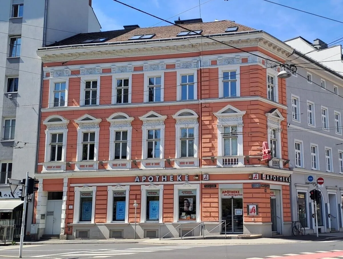 Lachsfarbene Gründerzeit-Fassade der Apotheke am Volksgarten mit Schriftzug „Apotheke“ und rotem Apotheken-A